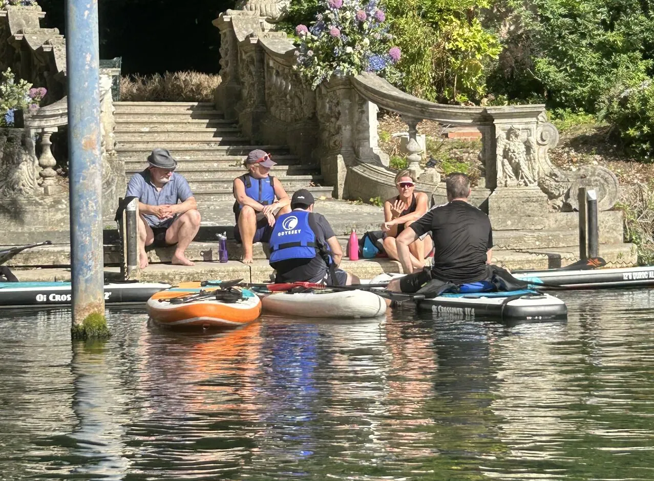 The objectsource senior leadership team sitting on paddleboards and steps on the bank of the River Thames, discussing the latest Adobe Commerce solutions.